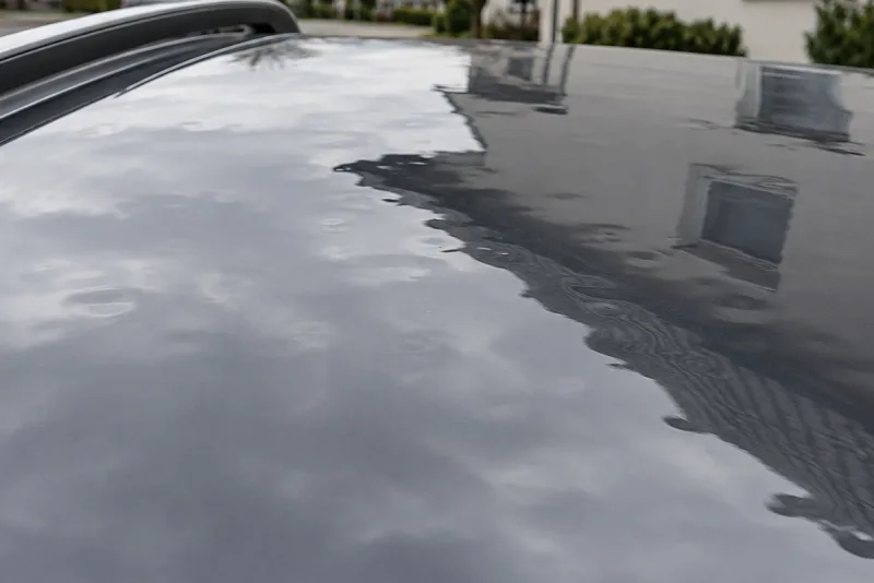 Close view across a dark grey car roof after a hailstorm: the reflected roofline and window frame break into small ripples at dozens of shallow dents.